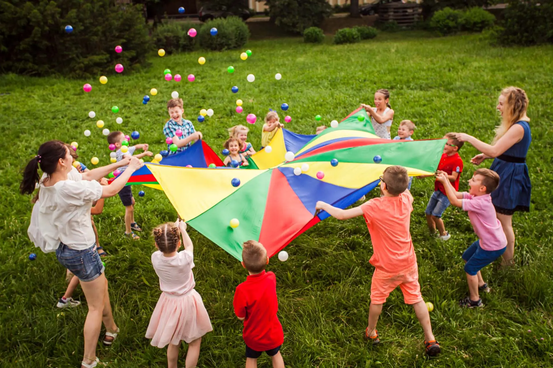 Happy kids waving rainbow parachute full of balls at green meadow